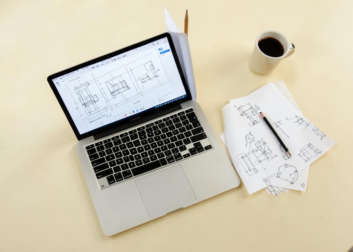 Laptop showing interface wireframes beside hand-drawn diagrams and a pencil on an ivory desk